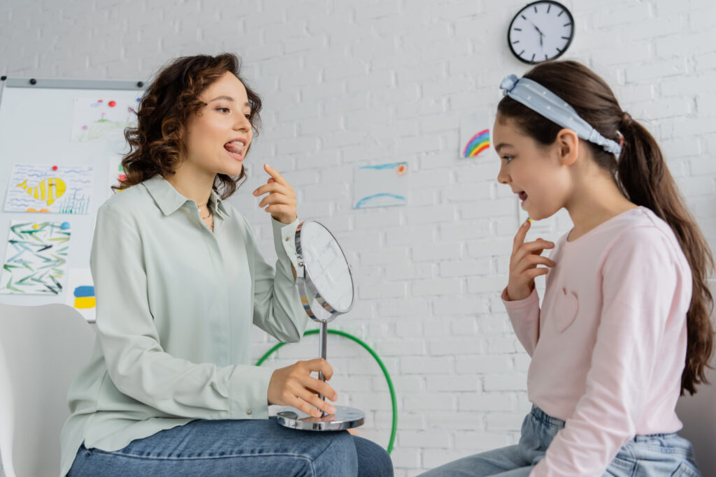 speech therapist holding mirror and pointing at mouth near pupil in consulting room